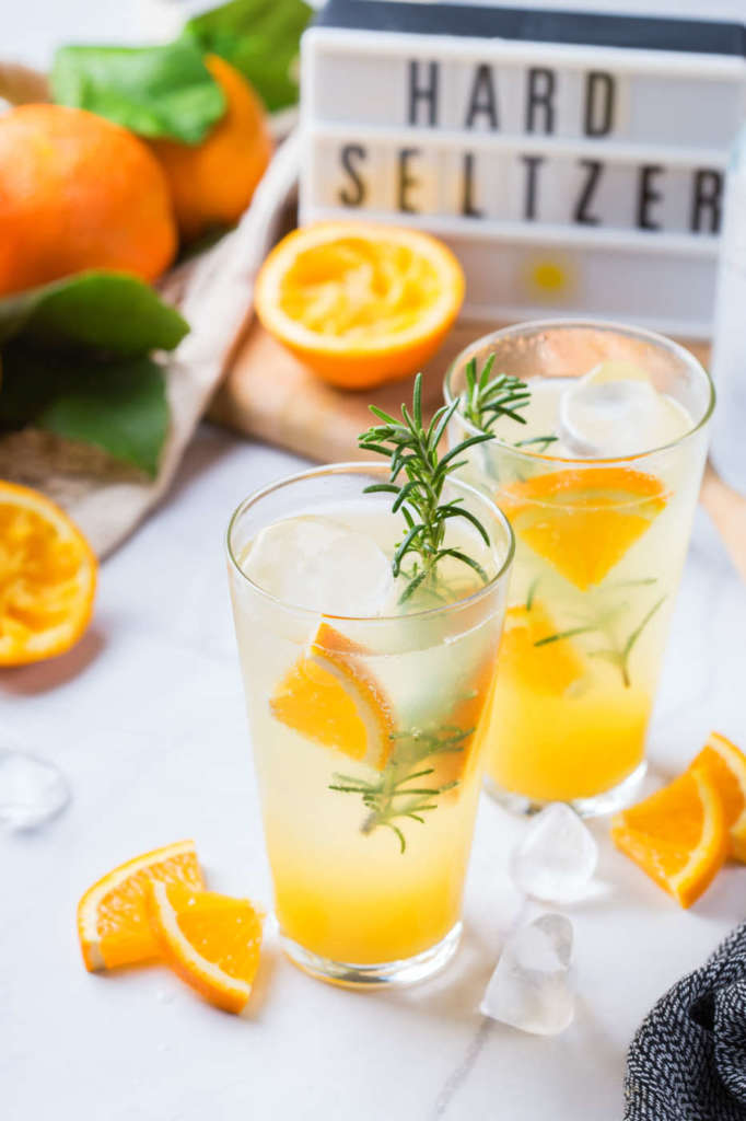 Hard seltzer cocktail with orange, rosemary and ice on a table. Summer refreshing beverage, drink on a white table